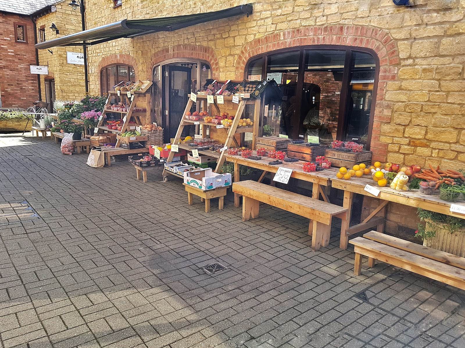The Harvest shopfront in the Fountain Court courtyard: honey stone walls, red brick arched windows and a green awning over wooden ladder shelves, benches and crates full of fruit and veg